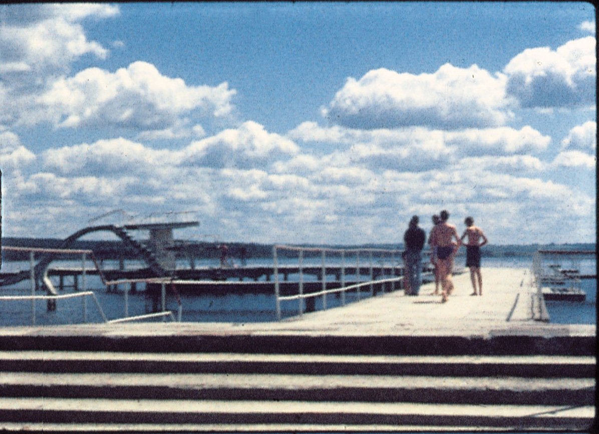 Narkevičius Foto eines Stegs, auf dem sich vier Personen, davon zwei in Badehosen befinden. Linkerhand gibt es eine Rutsche ins Wasser. Weiße Wolken sind am sonst blauen Himmel.