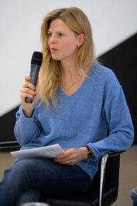 Foto von Lisa Moravec, einer sitzenden blonden Frau, die ein Mikrofon in der Hand hält und Zettel mit Text in der anderen.