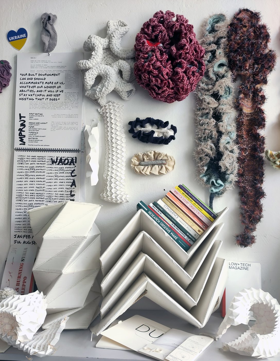 Unterschiedliche aus Textilien und Karton gefertigte Gegenstände sind nebeneinander drapiert und teils an der Wand aufgehängt. Auf gefaltetem Kartons ist ein Stapel Bücher. An der Wand hängt eine Drucksorte mit dem Titel "women and other anomalies" und ein herzförmiger Ukraine-Sticker.