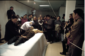 Foto der Performance Planting Grasses in der Fuck Off exhibition. Eine Person liegt auf auf dem Bauch auf einem Podest. Eine andere Person mit Maske und einem Gerät steht über den Rücken der liegenden Person gebeugt. Zuschauer stehen daneben, teilweise mit Kameras.