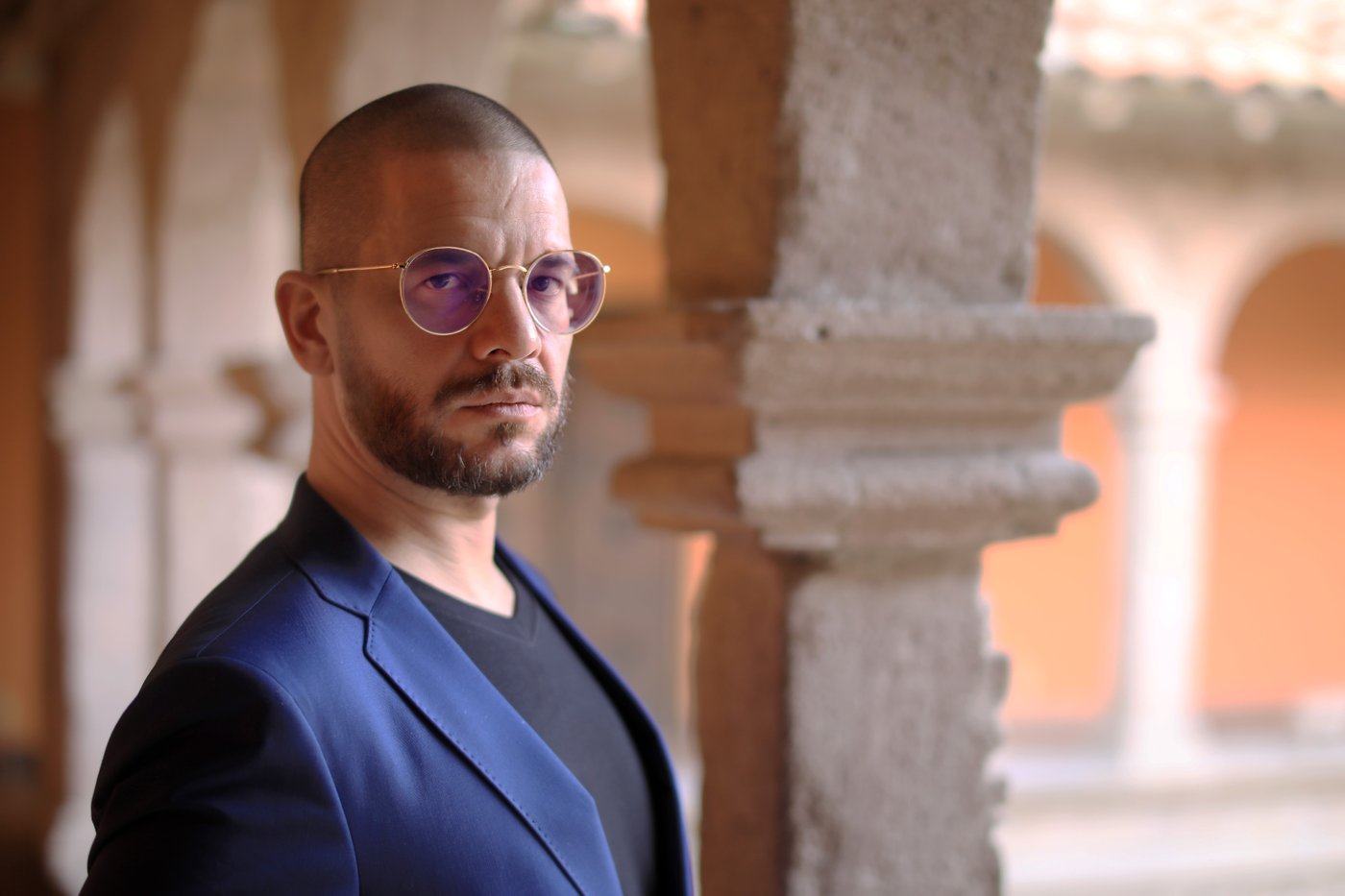 Foto einer Person im Halbprofil, mit dunklem Bart und Brille mit Goldrand. Im Hintergrund sind unscharf sandfarbene Säulen und Architektur zu erkennen.