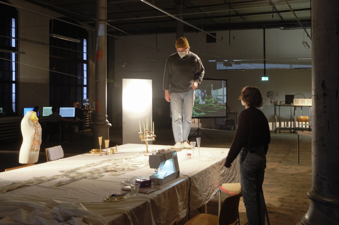 Raumgreifende Installation, Rundgang 2022, Foto © Lab_tf Ein Student steht auf einem mit einem Tuch gedeckten Tisch, auf dem eine Nähmaschine, ein Kerzenleuchter, goldenes Geschirr und diverse andere Utensilien liegen. Rechts daneben steht eine Studentin. Im Hintergrund sieht man den Atelierraum mit Screen, einem Modell und PC-Arbeitsplätzen.