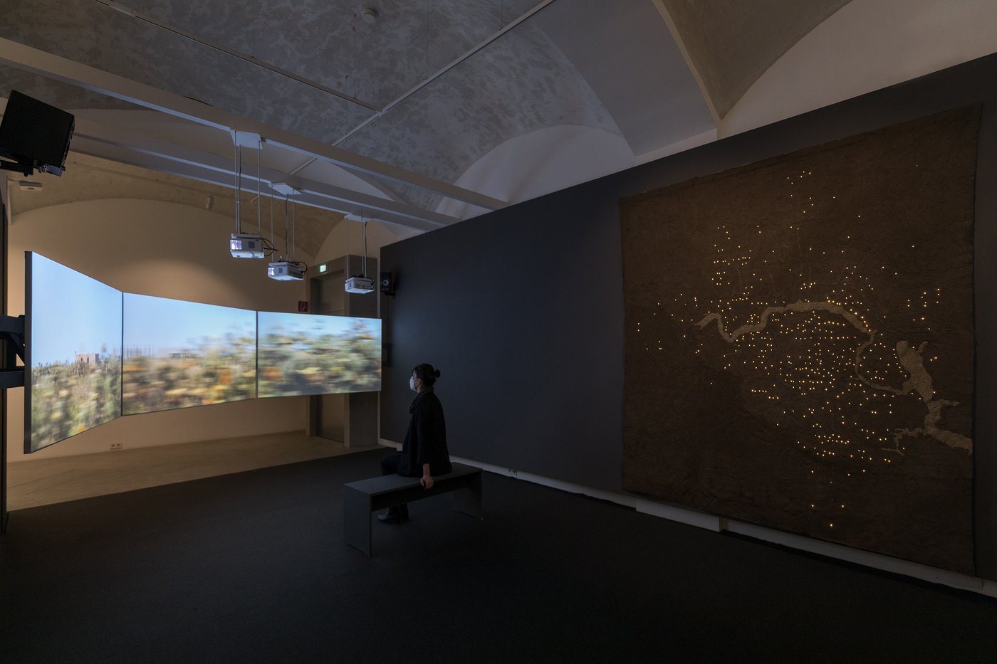 Ausstellungsansicht mit einer Besucherin, zu sehen ist eine Drei-Kanal-Videoinstallation auf der eine vorbeiziehende Landschaft gezeigt wird.