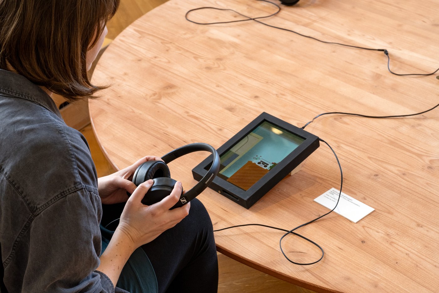 Person hält Kopfhörer in den Händen und blickt auf ein Tablet auf einem Tisch