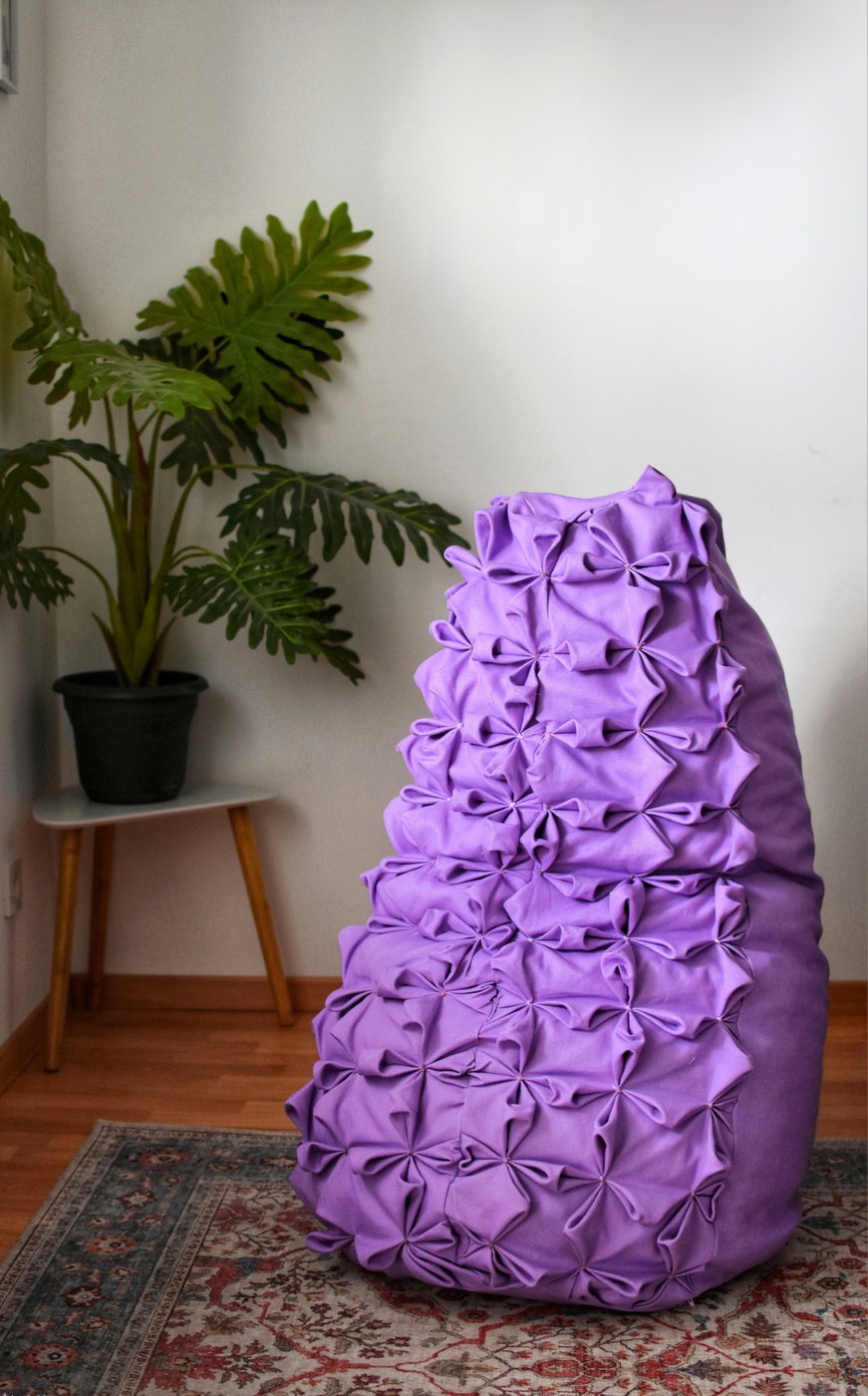 Ein lilafarbener Sitzsack, dessen Oberfläche mit aufwendiger Smockingtechnik gestaltet ist, steht auf einem Teppich. Daneben steht eine große grüne Zimmerpflanze auf einem kleinen Tisch vor einer weißen Wand.
A purple beanbag chair with an elaborate smocked surface stands on a rug. Next to it is a large green houseplant on a small table in front of a white wall.
