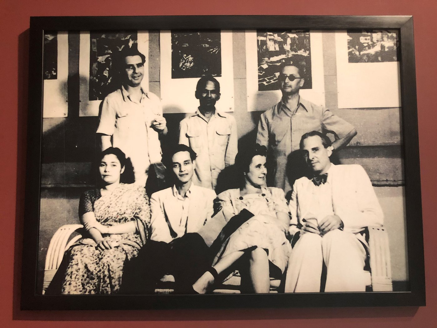 Die SW-Fotografie zeigt eine Gruppe von Künstler_innen vor einer Reihe von Grafiken, im Vordergrund auf einer Rattan-Bank sitzend: eine unbekannte Frau im Sari, Sayed Haider Raza und Käthe Langhammer mit ihrem Mann Rudi von Leyden. Dahinter stehen Walter Langhammer, K. H. Ara und Emanuel Schlesinger (mit Sonnenbrille).
