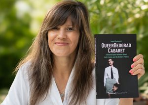 Die Farbfotografie im Querformat zeigt die lächelnde Autorin Ivana Marjanović. Das mittellange braune Haar trägt sie offen mit Scheitel auf der linken Seite, lange Fransen bedecken die Stirn. In der linken Hand hält sie das kürzlich erschienene Buch über das QueerBeograd Cabaret. Die Nägel sind lachsfarben lackiert. Im Hintergrund ist verschwommen Blattwerk zu erkennen.