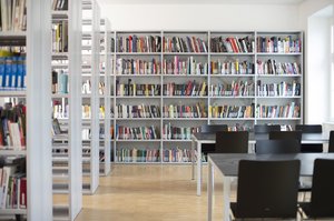 Die Farbfotografie zeigt einen lichtdurchfluteten Raum mit Holzboden und weißen Wänden. Links und an der Frontwand stehen mit Büchern befüllte Metallregale, rechts sind die Arbeitsplätze mit Tischen und Stühlen aus Metall und schwarzem Holz.