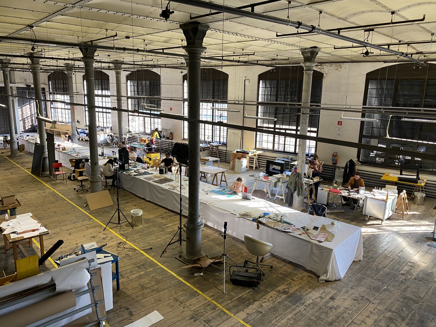 Installation in the studio, Open Days 2022, photo © Nina von Mechow A long table stands in a huge room with rows of columns and 6 large windows. Students are working at this and other workstations, work material is lying and standing around everywhere.