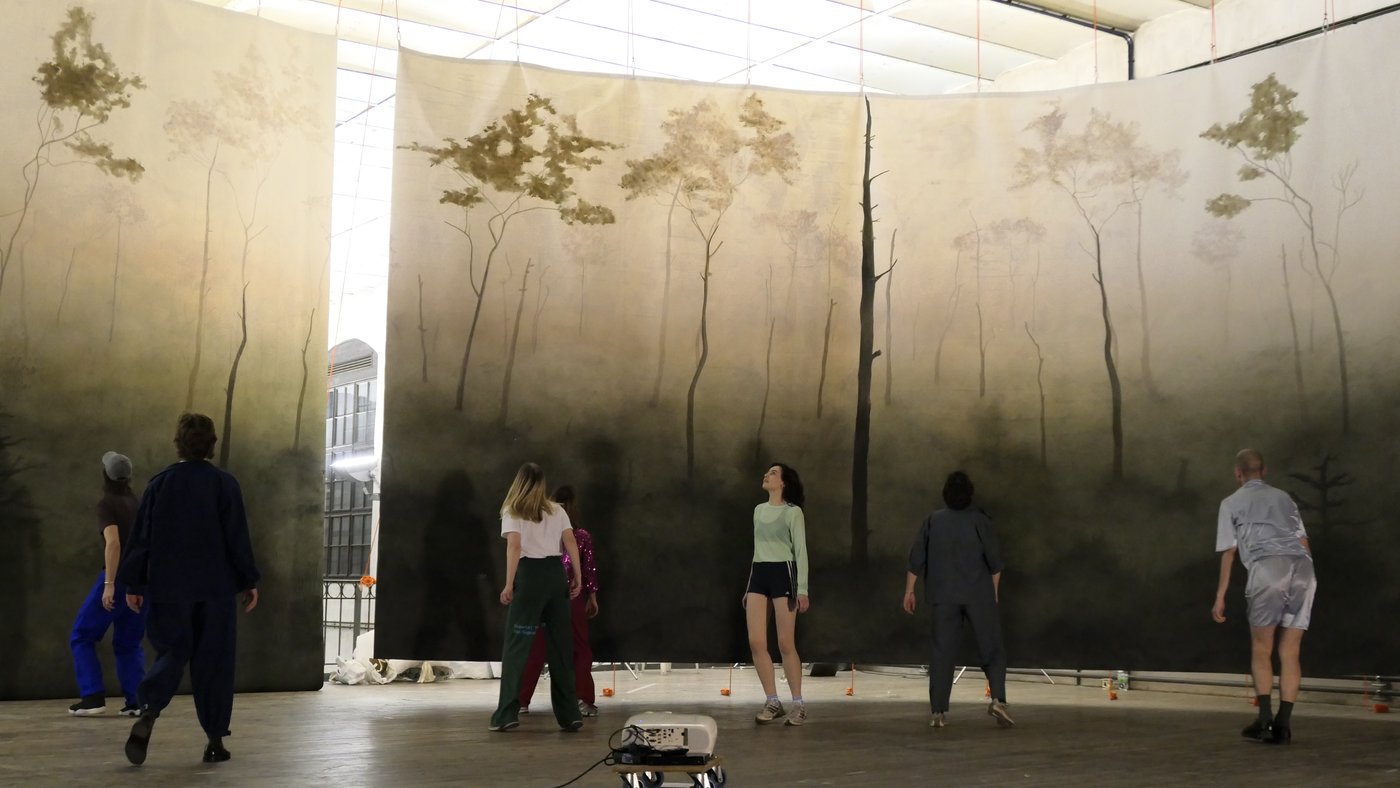 Rehearsal of the Diploma production, photo © Nina von Mechow In the background hang brochures painted with a light forest scenery, in front of them seven people obviously moving according to a choreography.