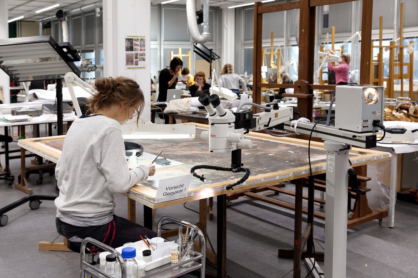 View of a large, bright studio room. On the table in the foreground is a canvas painting on which a female person is working. To the right is a microscope on wheels.