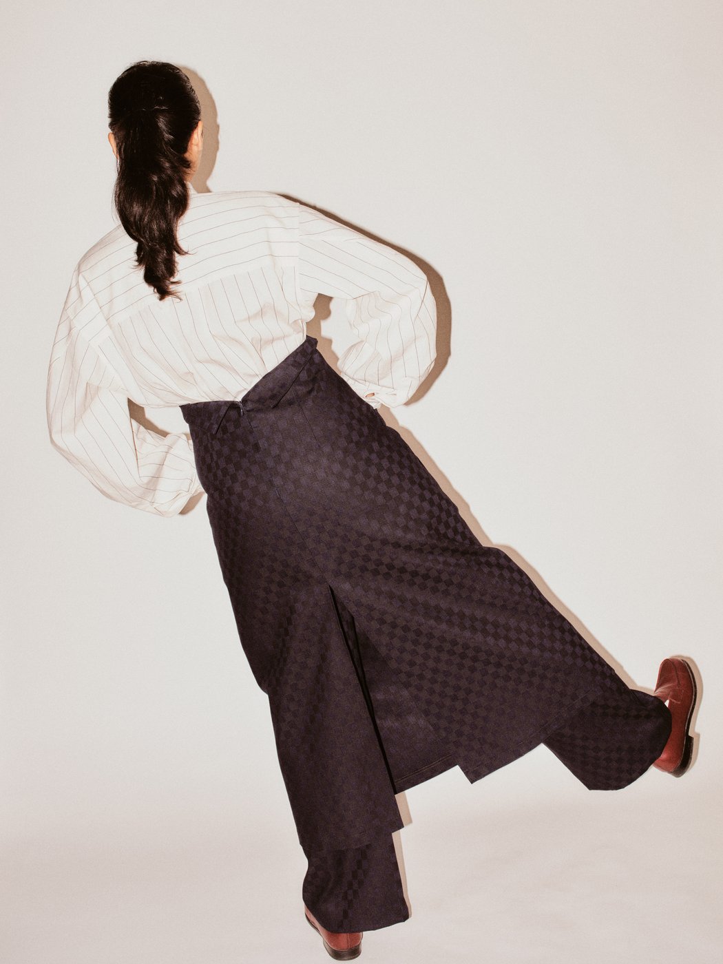 The image shows a person from behind. They are wearing a white shirt with fine stripes, wide dark checkered trousers, and a skirt with a long slit. One leg is raised, their hands are on their hips, and their hair is tied back.