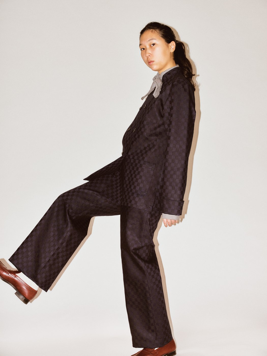 The image shows a person in front of a neutral background. They are wearing a dark checkered suit, a patterned shirt with a bow, and dark red shoes. The person is standing in a side pose with one leg bent and their hair tied back.