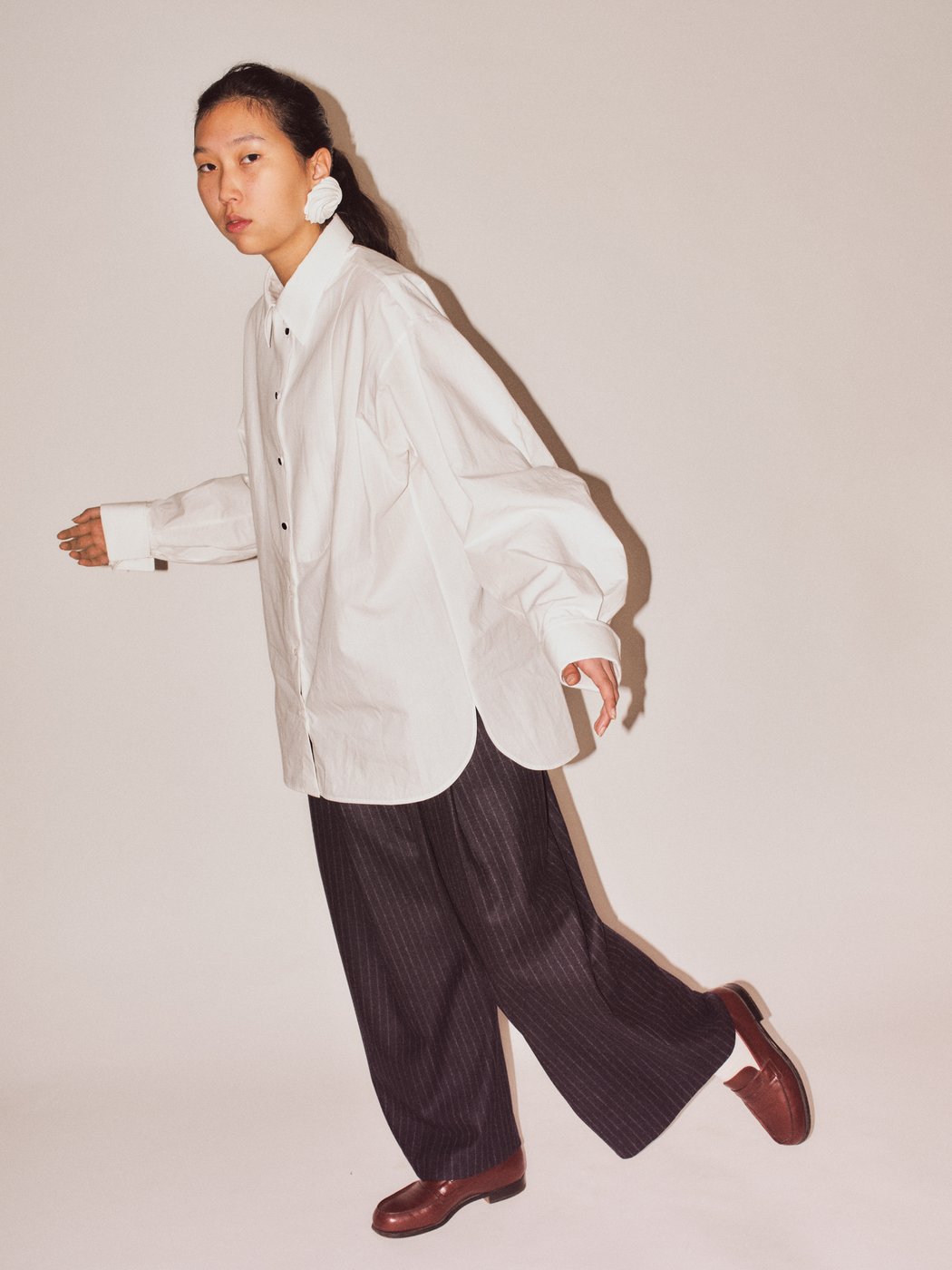 The image shows a person in motion. They are wearing a loose white shirt, dark pinstriped trousers, and dark red shoes. Their hair is tied back, and they are wearing striking earrings. The background is neutral.