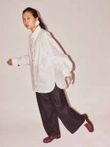 The image shows a person in motion. They are wearing a loose white shirt, dark pinstriped trousers, and dark red shoes. Their hair is tied back, and they are wearing striking earrings. The background is neutral.