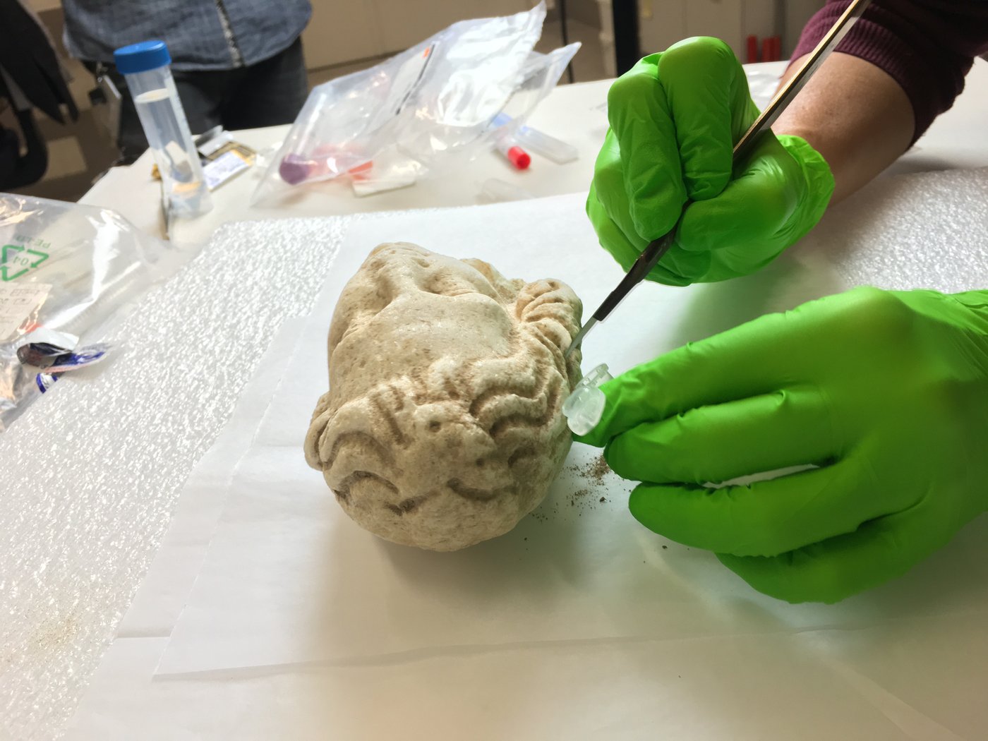 Gloved hands hold a scalpel to a small girl's head made of marble, below which is a sample tube that catches the scraped dirt particles.