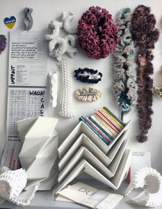Different items made from textiles and cardboard are placed next to each other and hung on the wall. A pile of books on folded cardboard. On the wall, there is a print titled "women and other anomalies" and a heart-shaped Ukraine sticker.