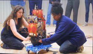 Photo of two young performers carrying a multi-masted model ship on blue fabric on wheels on the floor.