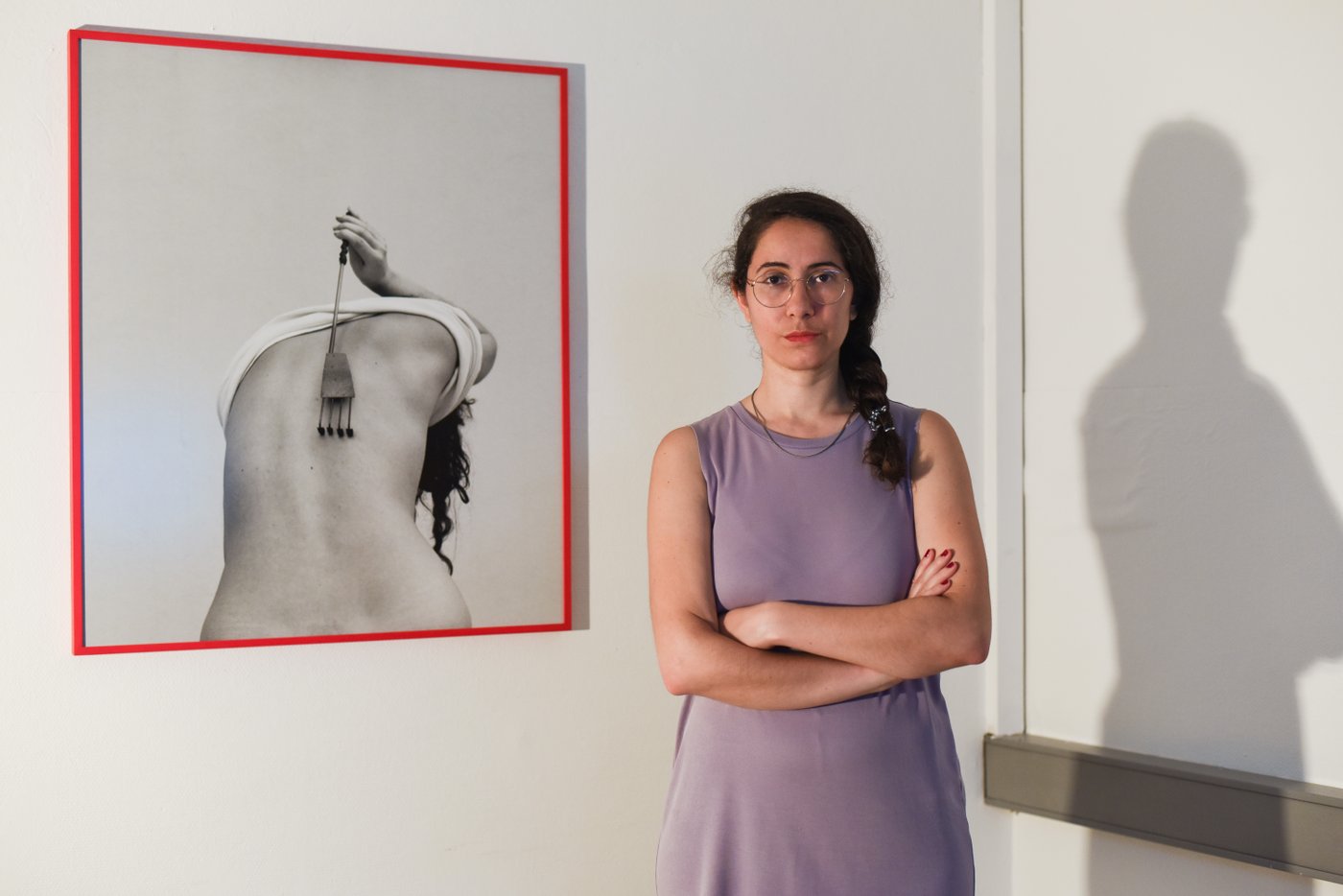 A person with dark hair in a lilac dress stands in a room in front of a photo of a person scratching his back with a fork.