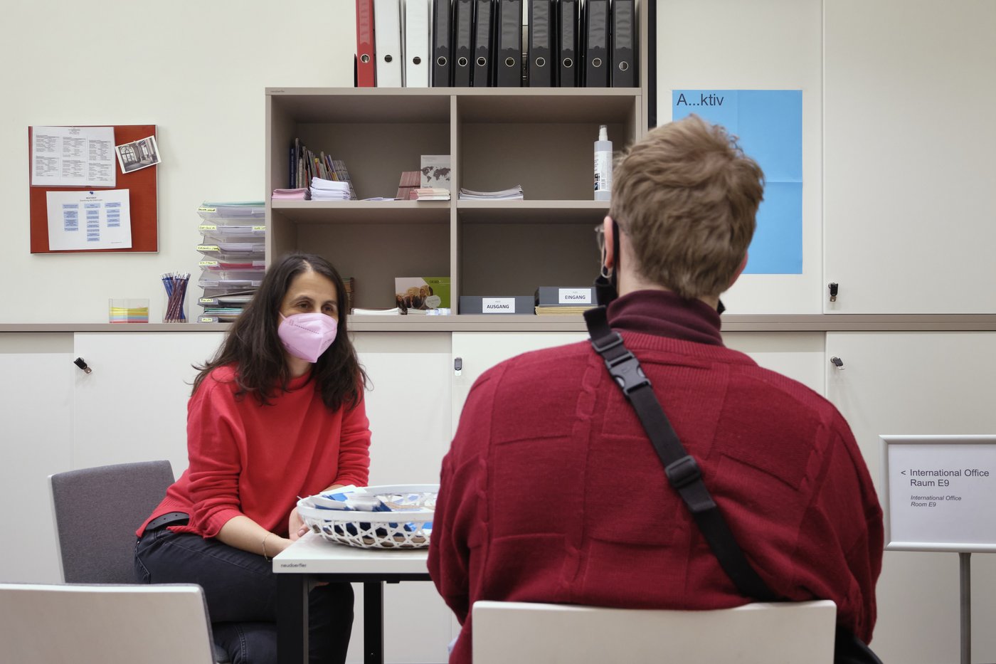 Consultation at the International Office, photo © Thomas Freiler A woman and a student are sitting at a table, both have red sweaters and masks on. In the background you can see typical office equipment.