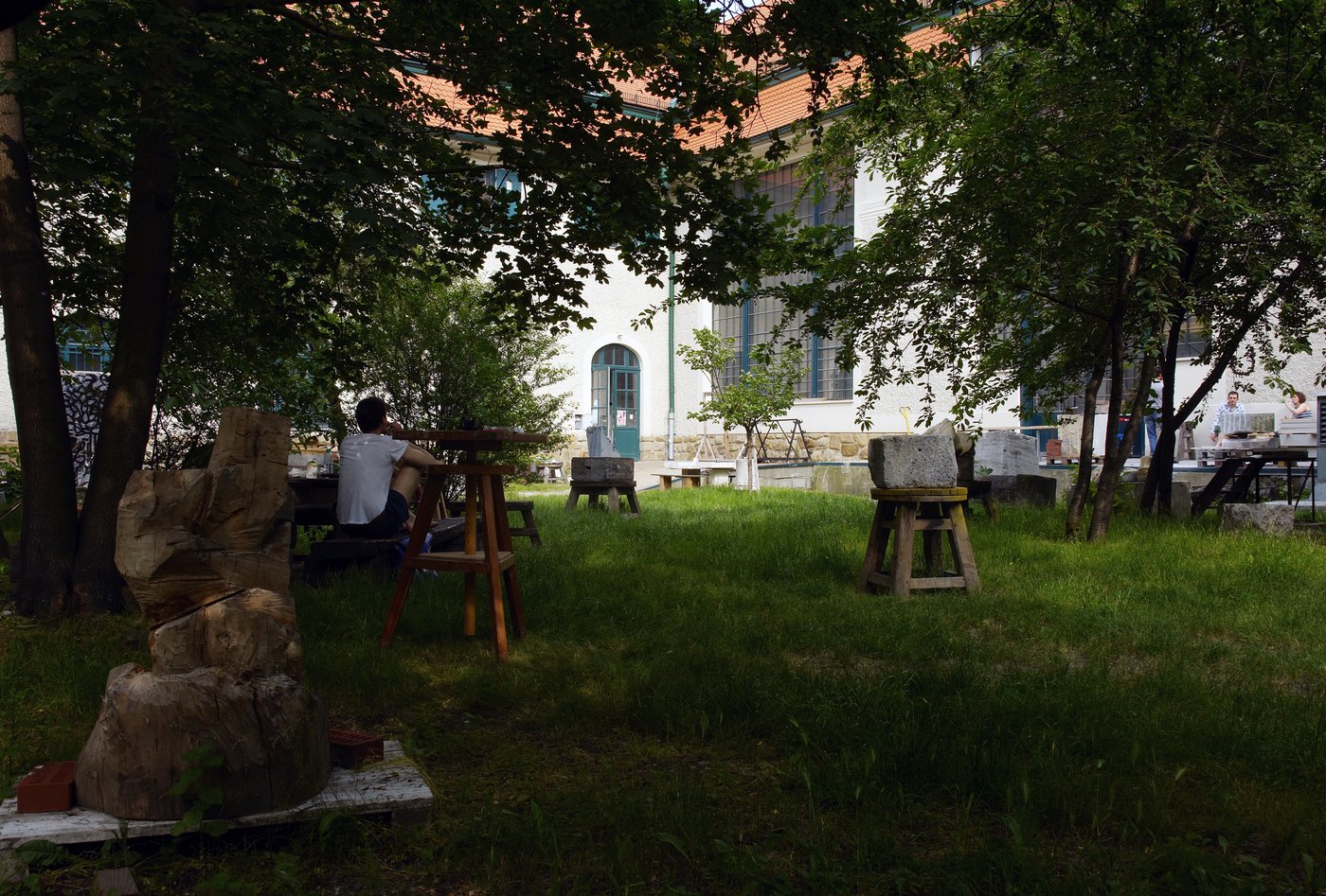 View into the garden of the sculpture studios, photo © Lisa Rastl In a summer garden, a young man sits with his back turned, his legs drawn up, wearing a white T-shirt. He is surrounded by wood and stone sculptures. In the background you can see the historic building with the large studio windows.